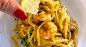 A hand with red nail varnish squeezes a lemon wedge over a plate of prawn pasta with yellow sauce, garnished with fresh herbs.