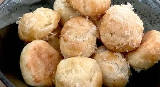 A dark bowl filled with round, golden-brown cheese puffs topped with grated cheese, sitting on a wooden surface.