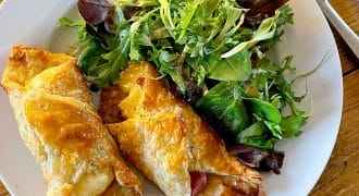 A white plate with two golden, flaky pastry rolls filled with cheese and meat, served alongside a fresh mixed green salad, set on a wooden table with a fork and knife.
