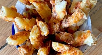 A bowl lined with greaseproof paper filled with golden, flaky cheese straws made from puff pastry, arranged upright on a wooden table.