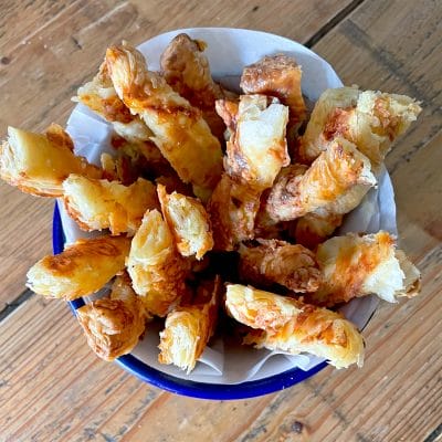 A bowl lined with greaseproof paper filled with golden, flaky cheese straws made from puff pastry, arranged upright on a wooden table.