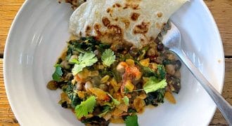 A white plate with chickpea and spinach curry garnished with coriander, served with two pieces of flatbread. A spoon rests on the plate, which is set on a wooden table.