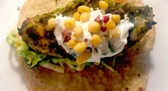 A wholemeal tortilla topped with grilled chicken, shredded lettuce, a dollop of soured cream, crispy round snacks, and red seasoning, served on a white plate.