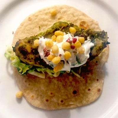 A wholemeal tortilla topped with grilled chicken, shredded lettuce, a dollop of soured cream, crispy round snacks, and red seasoning, served on a white plate.