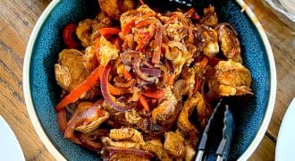 A bowl of cooked chicken strips with red onions and red peppers, served in a blue bowl with tongs. Grated cheese and tortillas are visible nearby on a wooden table.