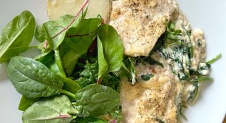 A white plate with creamy chicken and spinach, a halved boiled potato, and a fresh green salad with leafy greens.