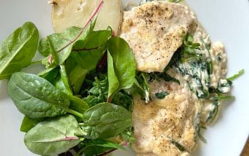 A white plate with creamy chicken and spinach, a halved boiled potato, and a fresh green salad with leafy greens.