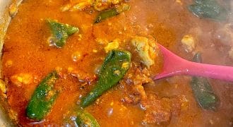A pot of red, spicy stew with chunks of meat and whole green peppers, being stirred with a pink spoon. Steam rises from the hot, simmering dish.