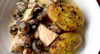 A white bowl contains creamy chicken and mushroom stew with herbs, meatballs, and two crispy, golden roasted smashed potatoes on the side.
