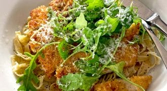 A white plate with pasta topped with crispy breaded chicken, fresh rocket, and grated cheese, with a fork and knife resting on the plate.