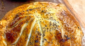 Golden-brown, round pastry topped with poppy seeds and decorative scored lines, resting on a baking tray. Another similar pastry is visible in the background.