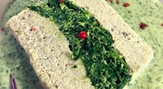 A rectangular slice of pâté with a green spinach centre is served on a creamy green herb sauce, garnished with pink peppercorns and small purple microgreens.
