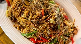 A large oval platter of stir-fried noodles with vegetables, chicken, and red and green peppers is placed on a wooden table, with serving tongs on each end of the dish. Steam rises from the hot food.