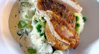 Sliced roasted chicken breast served over creamy pasta with garden peas and courgette, garnished with fresh herbs in a white bowl.