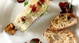 A gourmet dish presented on a white plate with two slices of crusty bread, a rectangular portion of food topped with microgreens and sauce, garnished with mushrooms and olives, and artistic sauce drizzles.