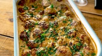 A baking dish filled with creamy meatballs garnished with chopped parsley, placed on a wooden table next to a glass of white wine and some drinking glasses.