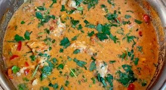 A metal pan filled with creamy, orange-coloured chicken stew, garnished with chopped fresh parsley and pieces of red pepper, sitting on a wooden surface.