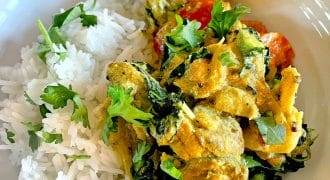 A white plate with steamed white rice, creamy yellow curry with chicken, spinach, tomatoes, and fresh coriander, garnished with a lemon wedge.