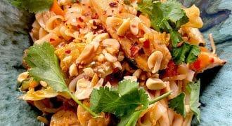 A close-up of a vibrant noodle dish topped with chopped peanuts, fresh coriander, red chilli flakes, and pieces of chicken, served in a green bowl.