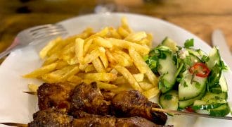 A plate with two grilled meat kebabs, a serving of golden chips, and a fresh cucumber salad garnished with herbs and red chilli slices. A fork and knife rest on the table beside the plate.