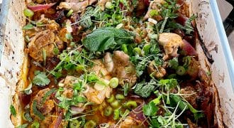 A baked dish filled with roasted chicken pieces, red onions, and herbs like coriander and mint, garnished with sliced spring onions and microgreens. The food is in a white rectangular baking dish on a hob.