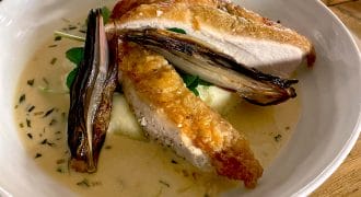 A white bowl with roasted chicken breast on creamy mashed potato, topped with grilled vegetables and a light herb sauce, served on a wooden table.