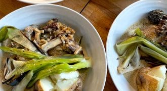 Four white bowls filled with baked potatoes, grilled spring onions, mushrooms, and creamy sauce are arranged on a wooden surface. The dish appears hearty and rustic, featuring roasted and sautéed vegetables.