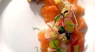Thin slices of raw salmon arranged on a white plate, garnished with microgreens, radish slices, cucumber, and a small pile of crumbled topping on the side.