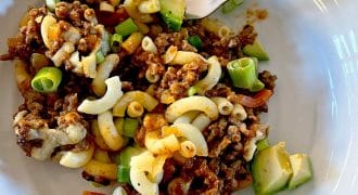 A white plate holds a serving of macaroni pasta mixed with minced beef, diced spring onions, avocado, tomato, and melted cheese. A fork rests on the edge of the plate.