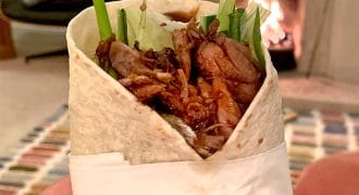 A hand with red nail varnish holds a wrap filled with shredded meat, lettuce, and cucumber. A fireplace burns in the background, creating a cosy indoor atmosphere.