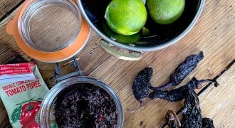 A bowl of three limes, a glass jar of dark paste, dried chillies, a tube of tomato purée, and an empty jar are arranged on a wooden surface.