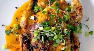 Grilled or roasted half chicken served on a white plate, topped with fresh microgreens, chopped spring onions, and herbs, with a pool of orange-brown sauce underneath.