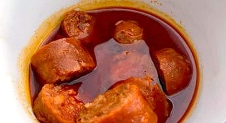 A white bowl with a blue rim contains several pieces of sausage in a rich, oily red sauce, likely a spicy or tomato-based stew.