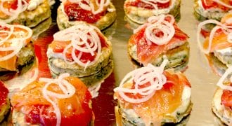 Rows of small, round appetisers on a metal tray, topped with cream, slices of smoked salmon, and thinly sliced onions. The appetisers are neatly arranged and garnished for serving.