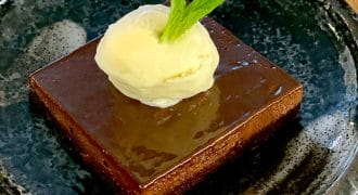 A square slice of chocolate cake topped with a scoop of vanilla ice cream and a fresh mint leaf, served on a dark ceramic plate with a rustic wooden background.