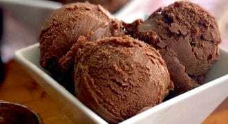 Three scoops of chocolate ice cream in a white square bowl, with more scoops and an ice cream scoop in the background on a wooden surface.