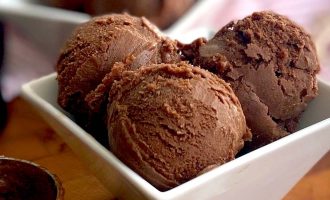 Three scoops of chocolate ice cream in a white square bowl, with more scoops and an ice cream scoop in the background on a wooden surface.