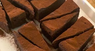 Slices of rich, chocolate cake dusted with cocoa powder are arranged neatly on a white rectangular plate on a wooden table.