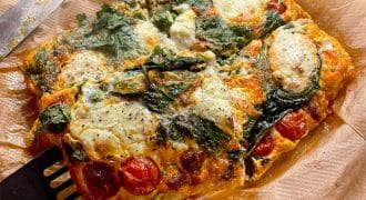 A rectangular baked frittata with spinach, cherry tomatoes, and melted cheese sits on baking parchment. A spatula is lifting a slice, and a knife rests beside it. The dish appears golden and freshly baked.