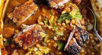 A pot filled with crispy roast pork belly pieces on top of a hearty stew with beans, sausage slices, chopped cabbage, and a rich, tomato-based broth. A spoon rests in the stew.
