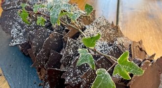 A chocolate yule log cake decorated with ivy leaves and dusted with icing sugar sits on a black slate platter atop a wooden table.