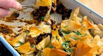 Hands serving a portion of a baked dish with crispy, golden filo pastry on top, garnished with chopped parsley, from a rectangular baking dish on a wooden table. Some filling and pastry remain in the dish.