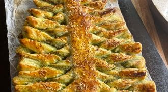 A baked puff pastry shaped like a Christmas tree, with twisted branches and sprinkled with sesame seeds, sits on baking parchment atop a wooden surface.