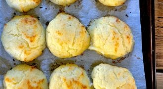 Nine golden-brown, fluffy baked scones are arranged on baking parchment in a baking tray. One scone is being lifted with a spatula, showing its light, airy texture.