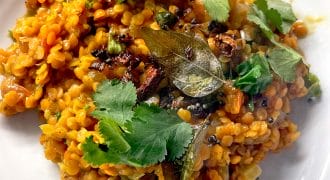 A serving of orange lentil dahl garnished with fresh coriander leaves and bay leaves, plated on a white dish. The dahl appears creamy with visible spices and herbs mixed in.