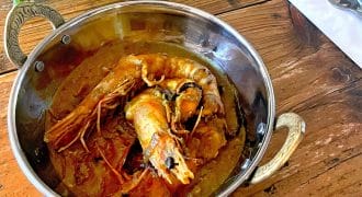 A metal bowl with ornate handles holds a prawn curry in a rich, reddish sauce. The bowl sits on a rustic wooden table beside a folded serviette with a fork and spoon, and some fresh herbs are visible nearby.
