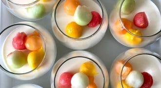 Several glasses filled with a creamy white dessert are topped with colourful melon balls in shades of orange, yellow, red, and green, arranged neatly on a white surface.