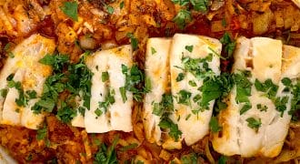 A baked dish featuring four pieces of white fish fillet on a bed of seasoned orzo pasta, topped with chopped fresh herbs, all served in a white oval baking dish.
