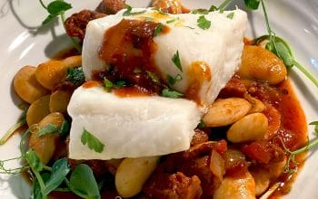 A dish featuring a piece of white fish atop a stew of large white beans, tomato-based sauce, and herbs, garnished with fresh greens on a white plate.