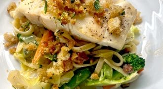 A piece of white fish topped with crispy breadcrumbs sits on a bed of mixed vegetables and pasta, served on a white plate.
